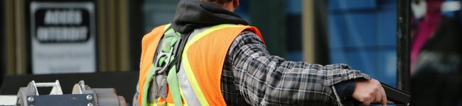 A construction worker wearing a blue helmet and safety vest operates equipment at an urban construction site.
